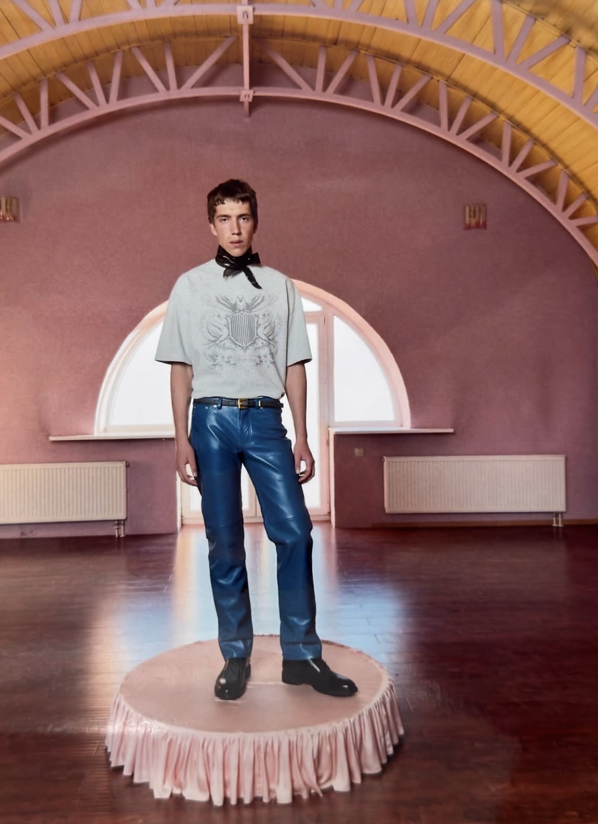 Model on a round pink platform wearing a gray crest-print T-shirt, black neck scarf and shiny blue trousers.