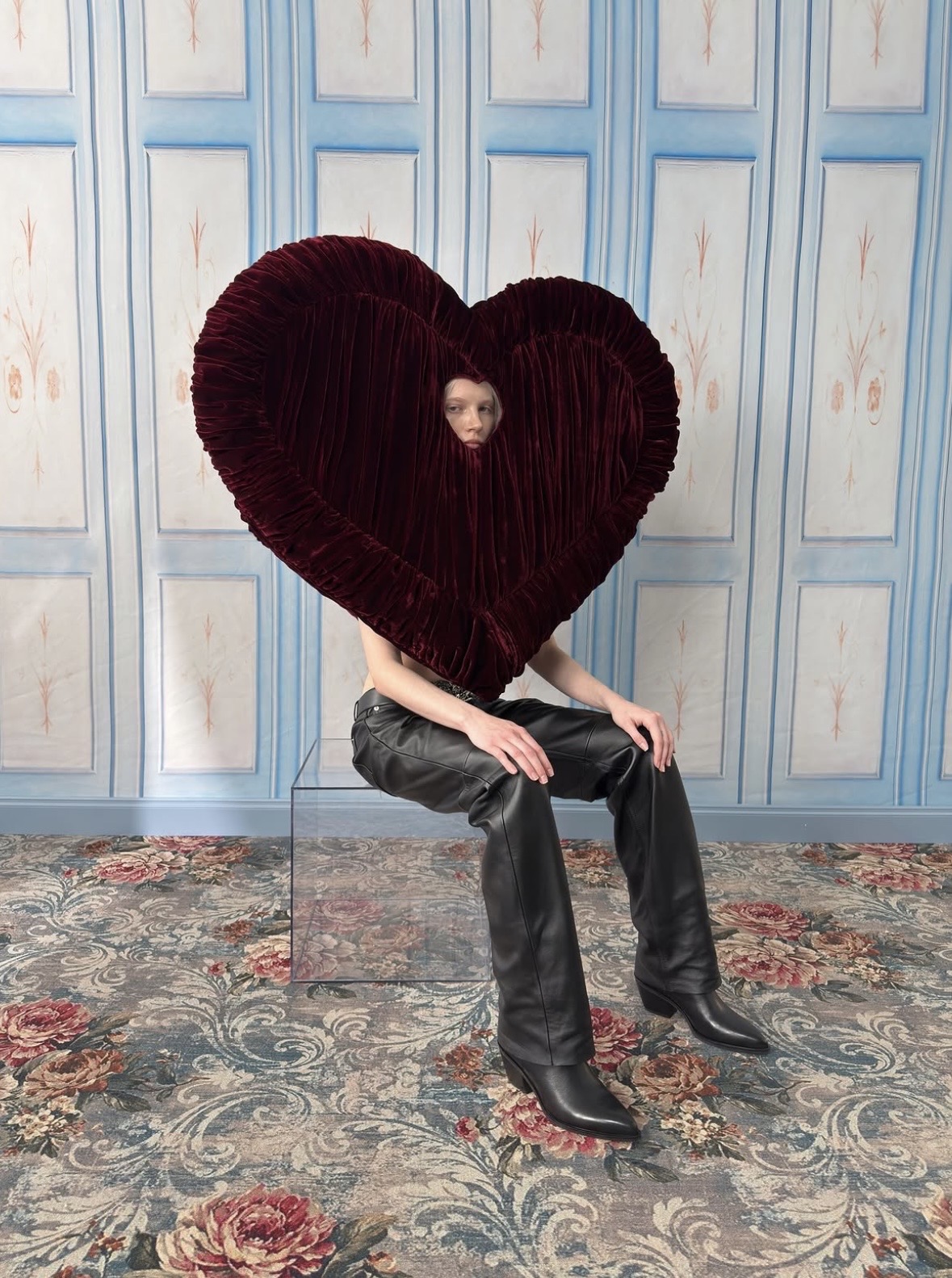 Seated model whose face is framed by a huge burgundy pleated heart, with black trousers and pointed black boots.