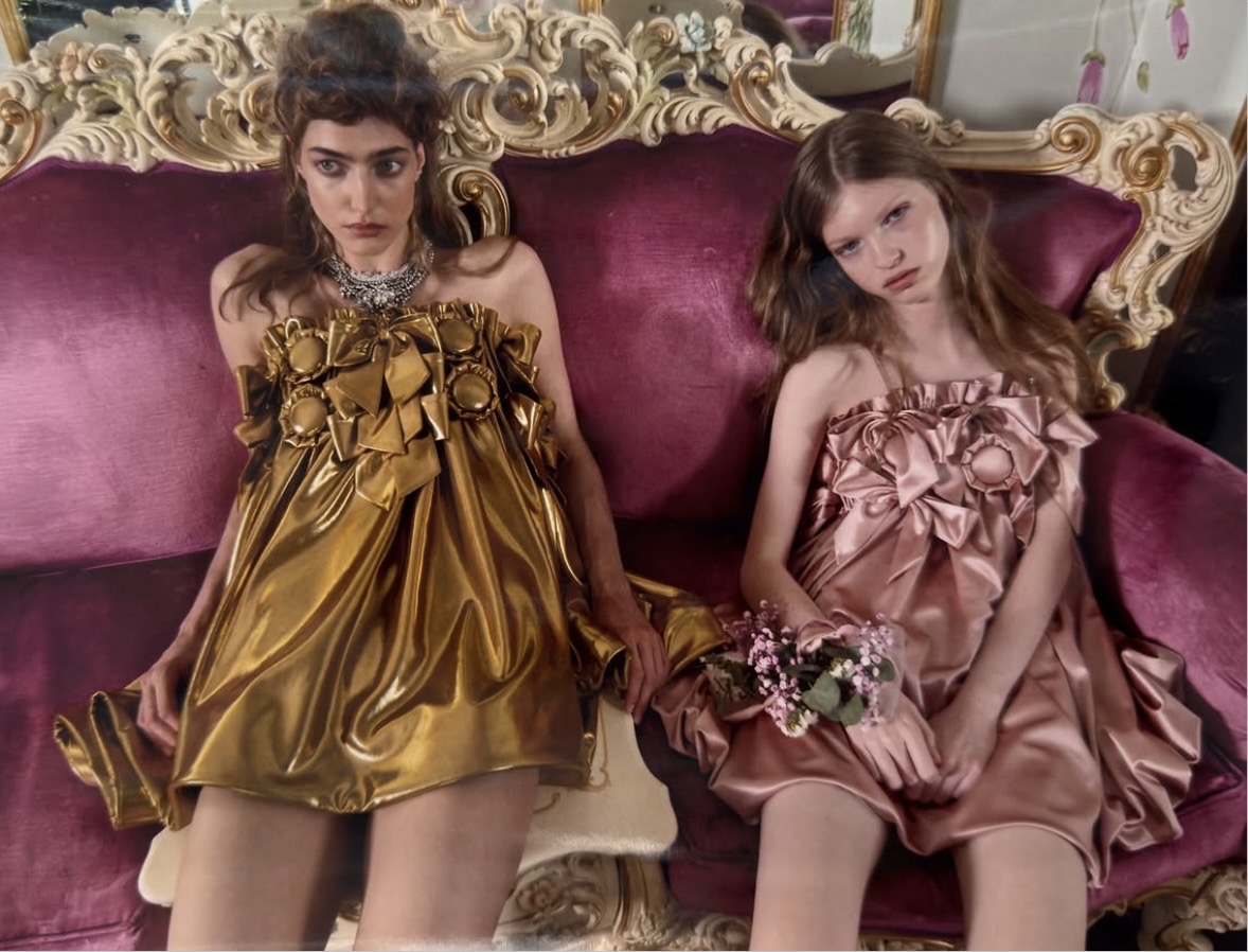 Two models seated on a pink-and-gold sofa in bronze and pale-pink mini dresses with oversized floral embellishments.