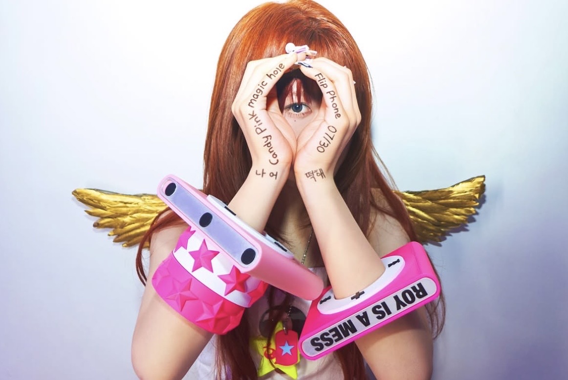 Red-haired model with gold wings framing one eye with her hands, wearing pink iPod bangles and star bracelets.