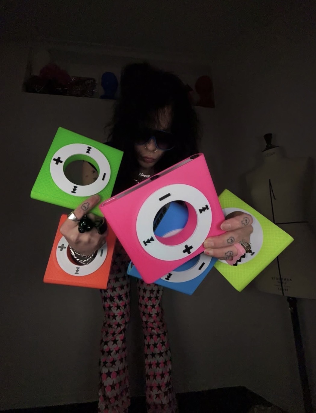 Dark portrait of a person leaning forward with oversized neon iPod-shaped bangles held toward the camera.