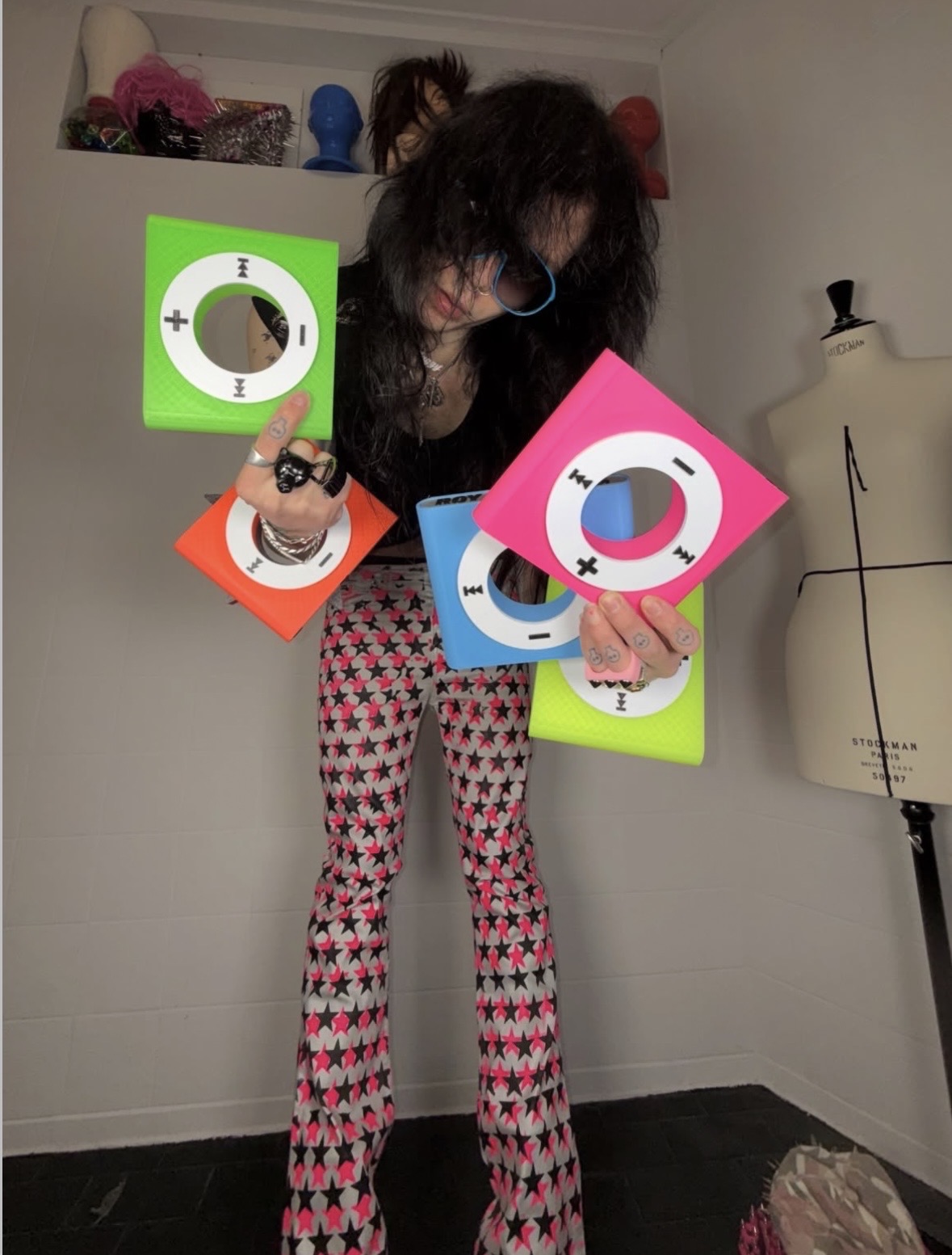 Standing portrait of a person displaying green, pink, orange and blue iPod bangles over patterned flared pants.