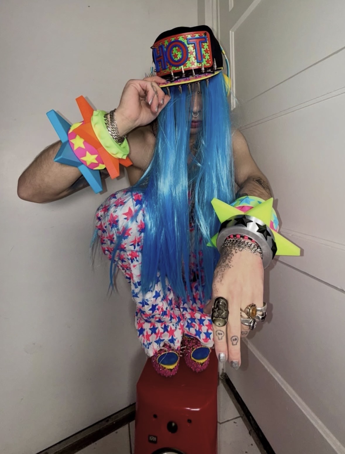 Blue-haired person crouching in a colorful HOT cap, star-print pants and stacked neon star bangles.