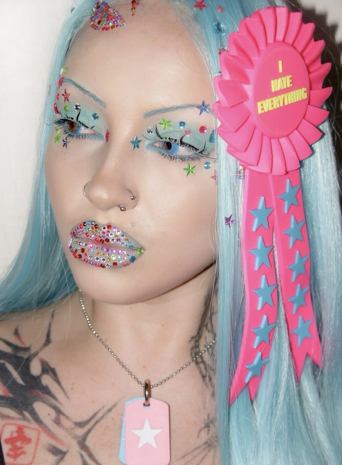 Blue-haired portrait with multicolored face gems, a star pendant and a large pink I HATE EVERYTHING rosette.