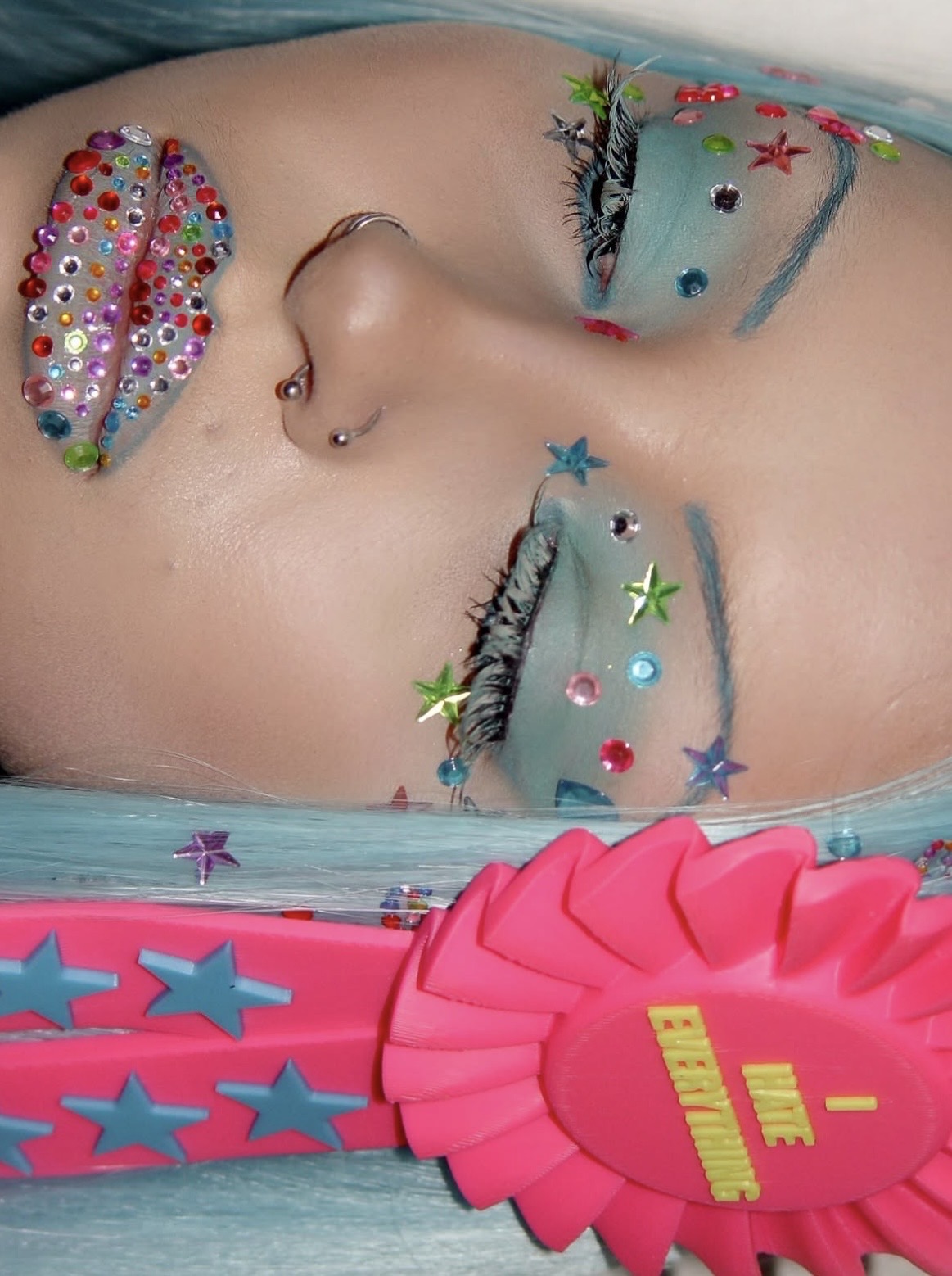 Sideways close-up of jeweled lips, star-covered eye makeup and a pink rosette against pale blue hair.