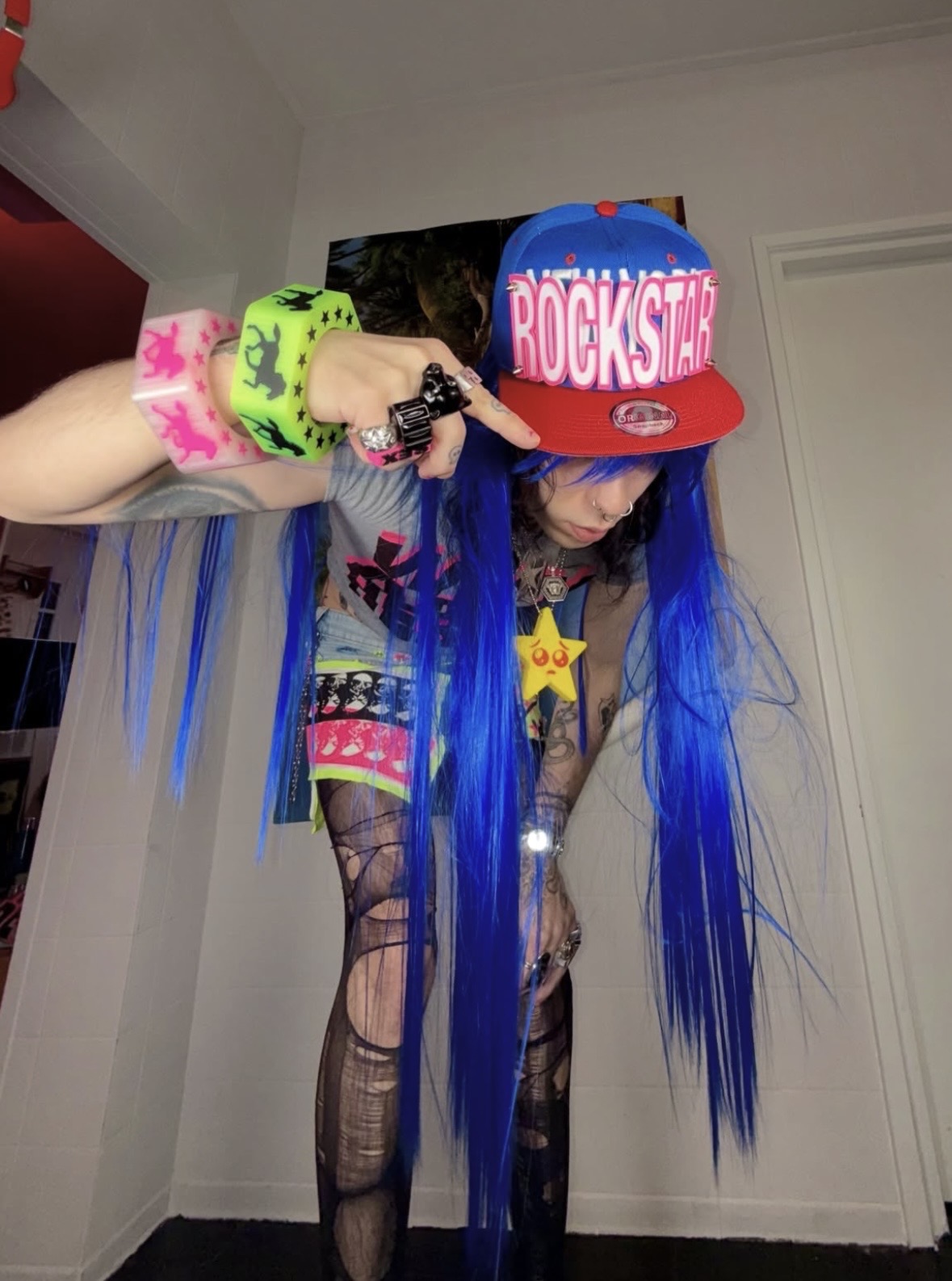 Person with long blue hair bending toward the camera in a ROCKSTAR cap and neon wrist cuffs.