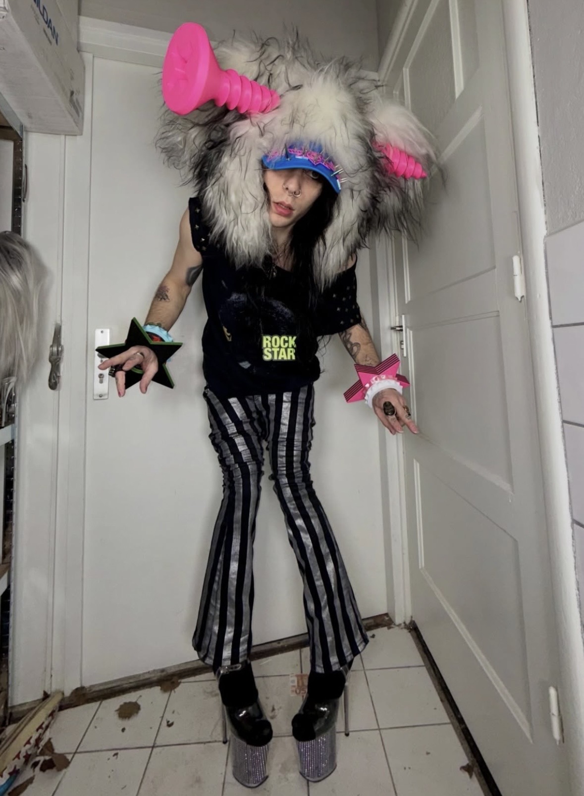 Person in striped pants and a ROCK STAR top wearing a shaggy headpiece with giant pink screws and star bangles.