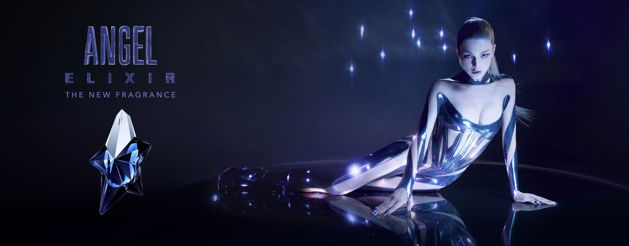 Hunter Schafer in a reflective outfit beside a star-shaped perfume bottle in Mugler's blue-lit Angel Elixir campaign.