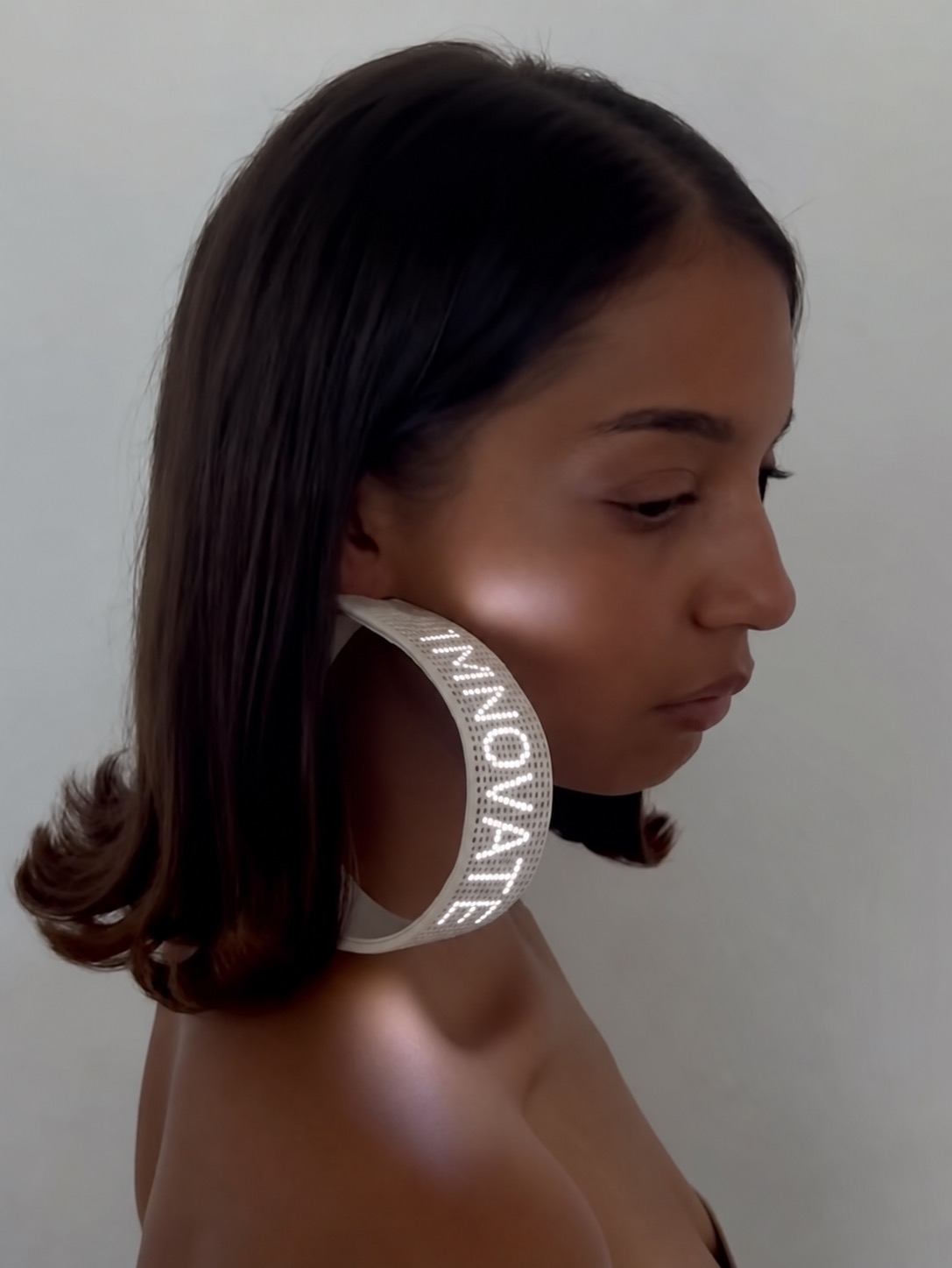 A side portrait wearing a sculptural screen earring with vertical Monamobile text.